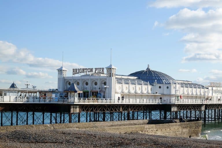 Exterior view of the Palace Pier in Brighton, East Sussex, U.K.  The large white building has a sign over the entrance and a large dome in the center.  The blue sky has a few white, fluffy clouds, and the base of the pier is visible under the building.
